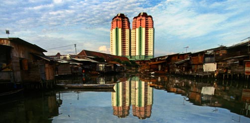 INDONESIA Jakarta. Man in a wooden boat navigating a polluted waterway backed by wooden slum housing, in contrast to the high rise appartment buildings visible behind.
Credit: Mark Henley / Panos.