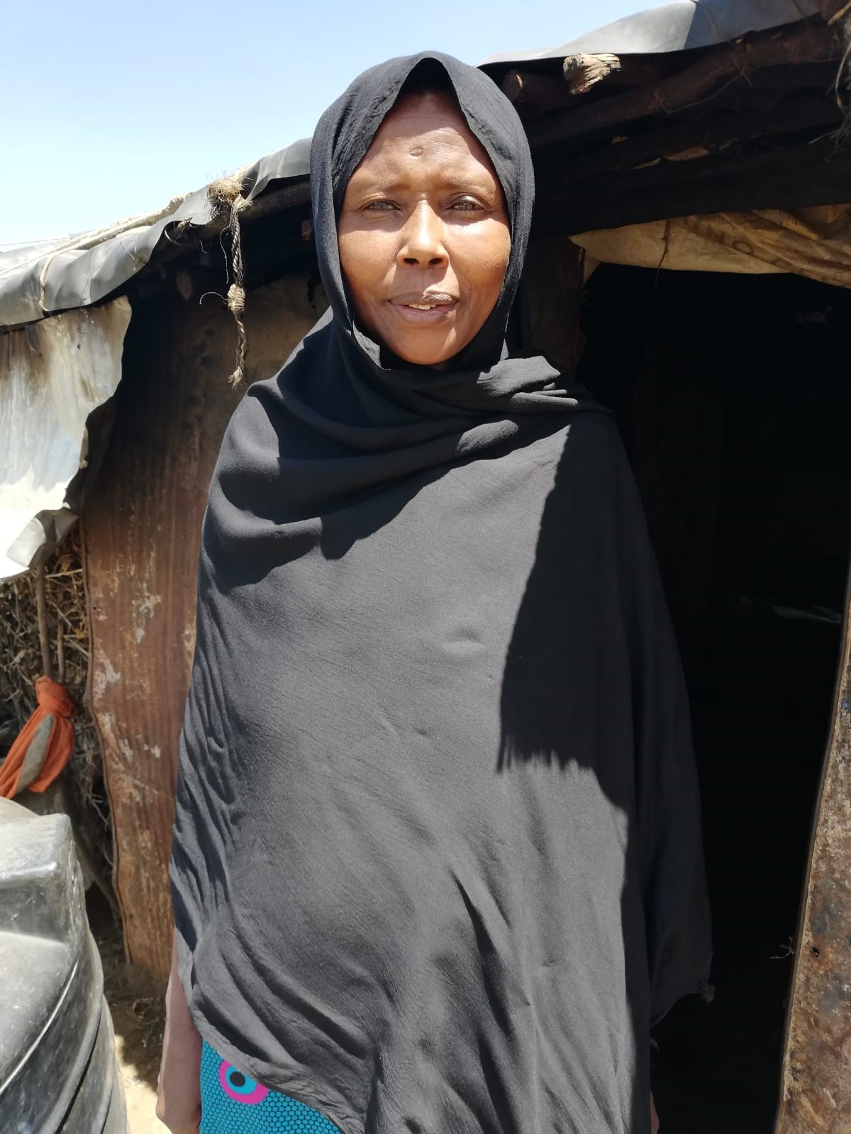 Fatuma stands looking at the camera, in front of her house. 