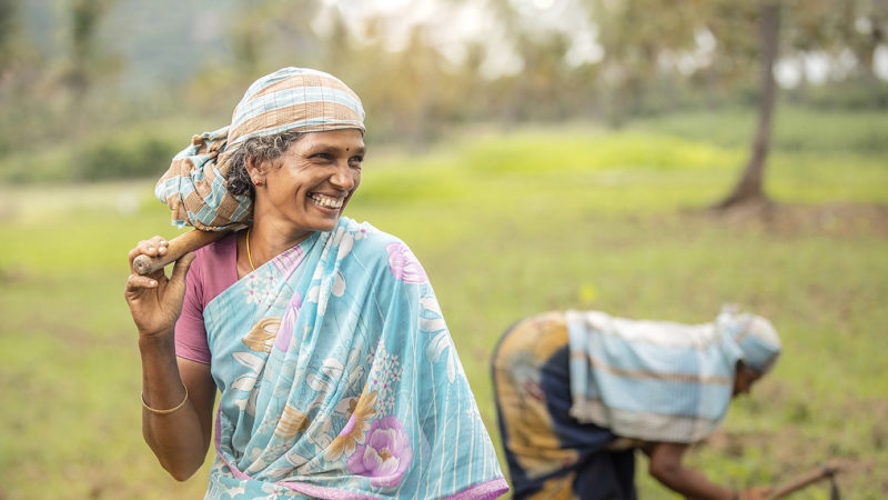 Woman smiling and holding a hoe over her shoulder while working in a field in Tamilnadu, India.