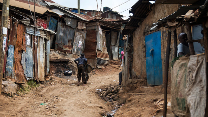 A man pushing a wheelbarrow through a red dirt street with shacks in Kibera - an informal settlement in Nairobi, Kenya.