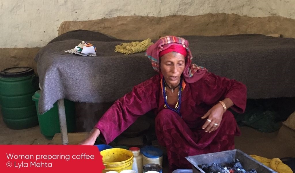 A woman preparing coffee on the floor of her humble home with many plastic containers around her