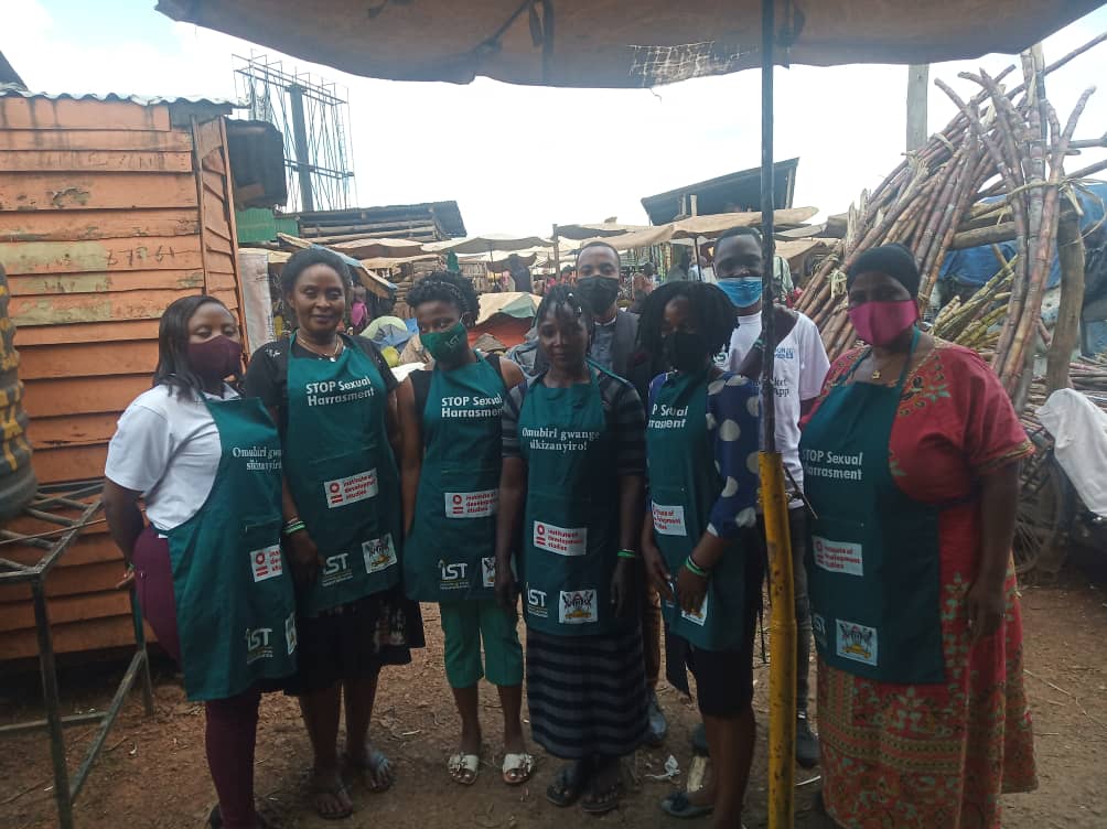 Five women and two men are in a market, wearing face masks. The women are wearing aprons that read 'stop sexual harassment' and 'my body is not a playground'.