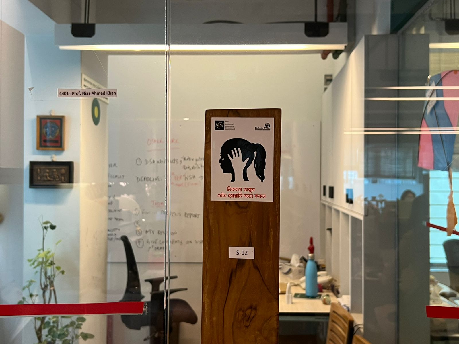 An office environment showing a chair, whiteboard, desk. On the wall is a poster with the outline of a woman's head. Within the outline is a hand and a tear.