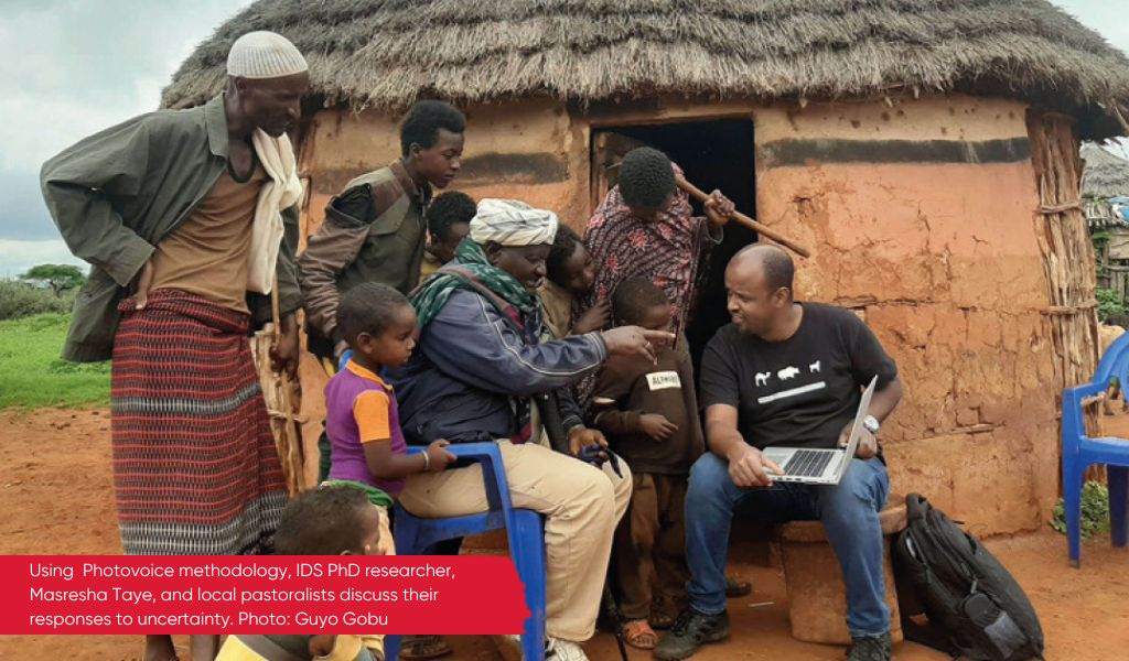 In Ethiopia, a man is sitting on a chair in front of a mud hut with a laptop on his lap. He is showing a group of pastoralists what is on his screen. They are discussing their responses to uncertainty.