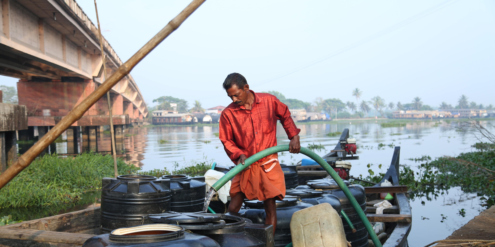 In Kuttanad in Kerala, India, a man is standing on a boat holding a green hose and is filling drinking water into water tanks to transport them by boat to different locations in the region. He is wearing a red shirt and shorts and there is a river behind him. A bride croses the river on the left of the photo.
