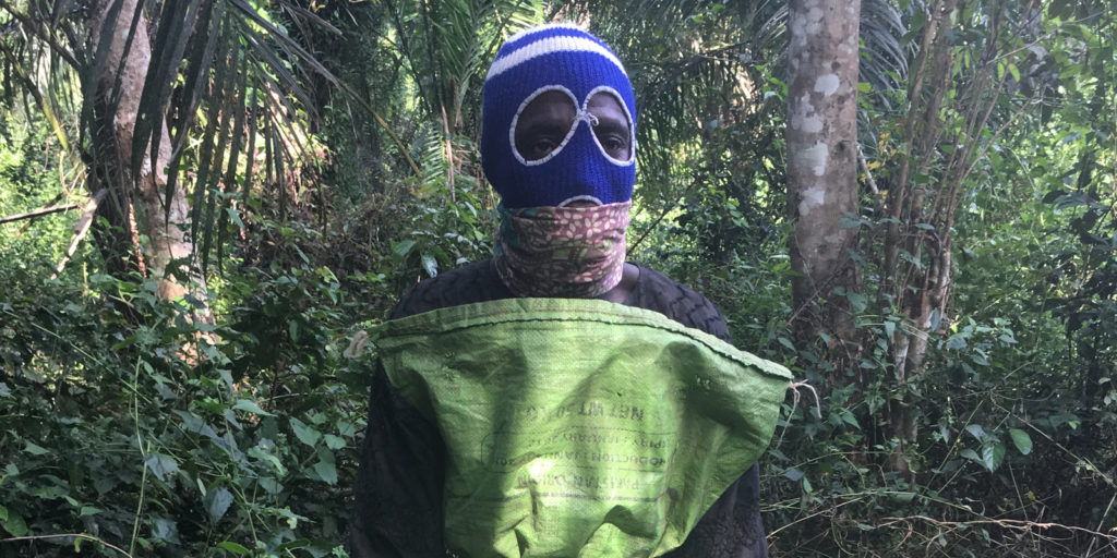 A man stands against a backdrop of a forest wearing a blue balaclava and a face mask.