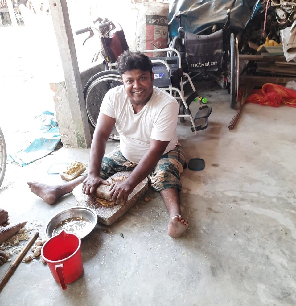 A man sitting on the floor, grinding spices and smiling, with his empty wheelchair behind him