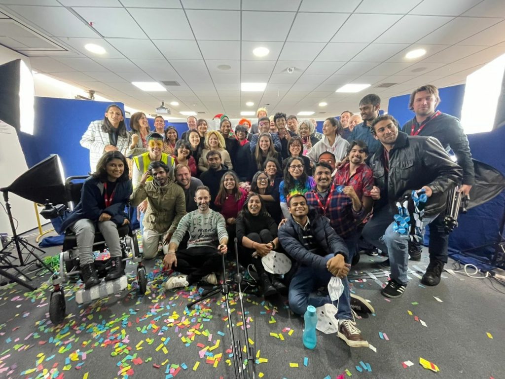 A group shot of dozens of IDS staff and students taken after the IDS panto performance. There is colourful confetti on the floor and stage lights on the right sna left. Everyone is looking at the camera and smiling.