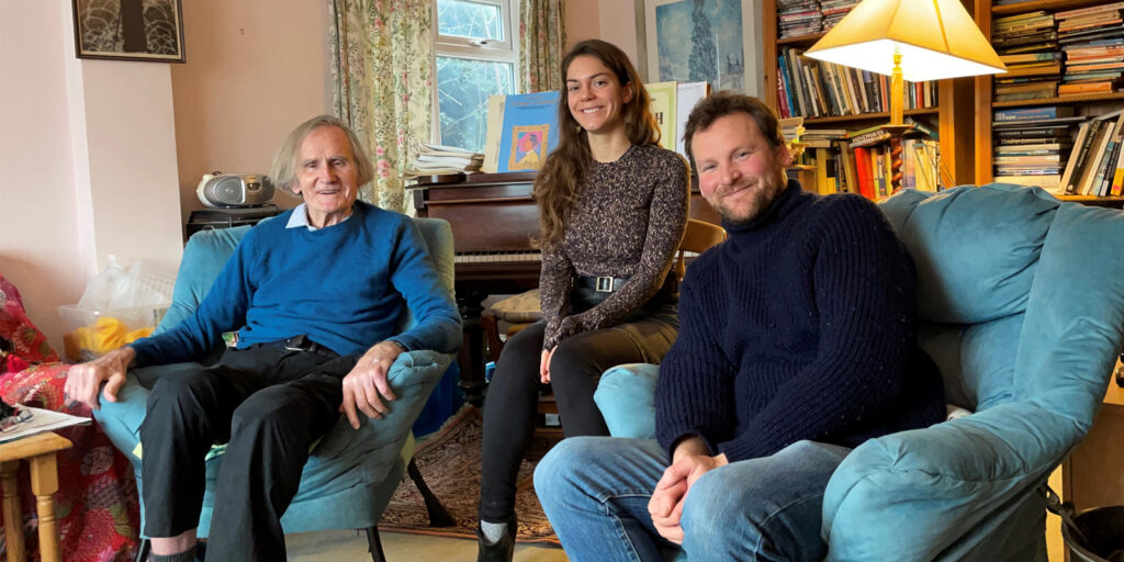 Robert Chambers sits on a chair next to Mariah Cannon and Stephen Thompson. They are in Robert's front room