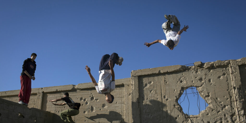 A photo of two male youths upside-down in the air after jumping from a wall in a cemetery on the outskirts of their refugee camp in Khan Younis, Gaza. The wall behind them shows damage from past Israeli incursions.
