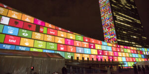 A photo of the United Nations building lit up with the colours and logos of the Sustainable Development Goals (SDGs).