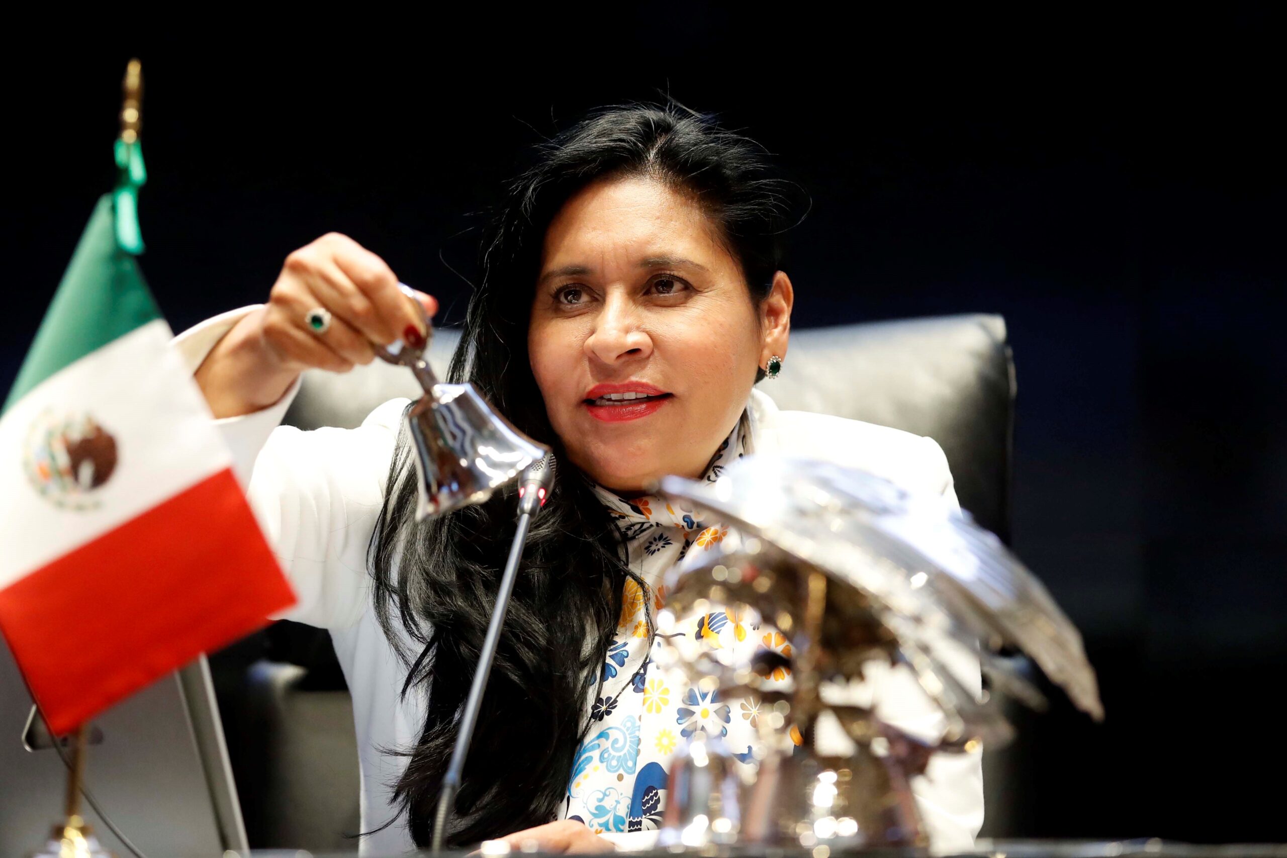 Portrait of a woman ringing a small bell, with a Mexican flag in the foreground.
