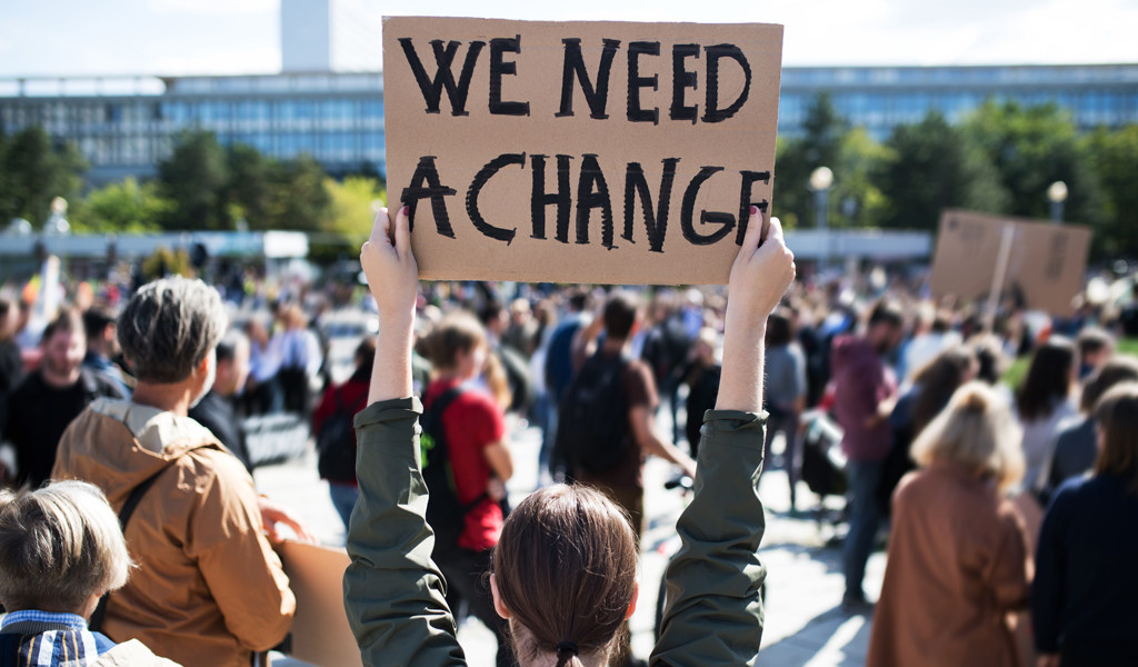 A photo of rear view of people with placards and posters on global strike for climate change. The main placard read: We Need a Change.