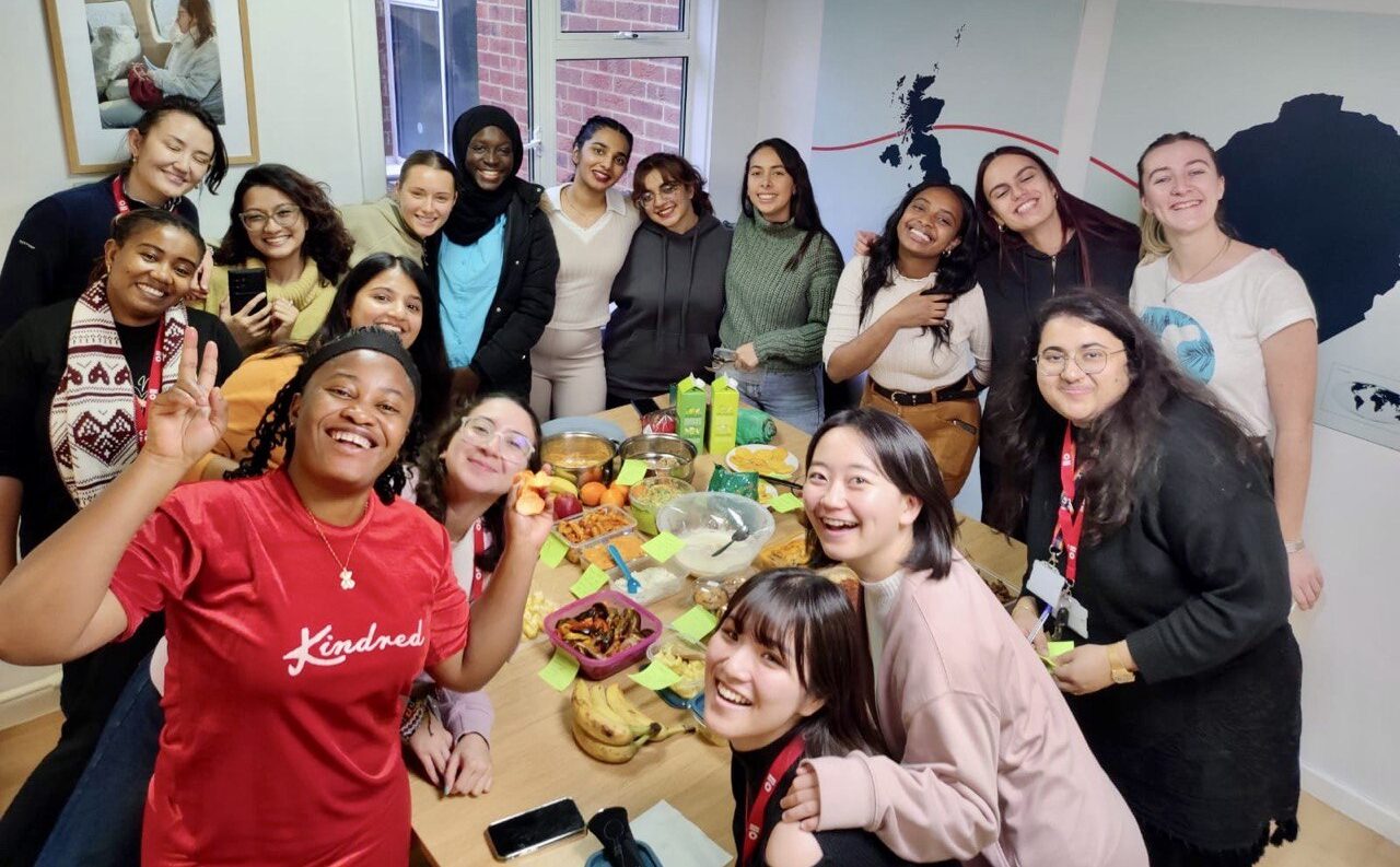 a large group of people are stading together around a table looking at the camera and smiling. On the table are plates of food to share.