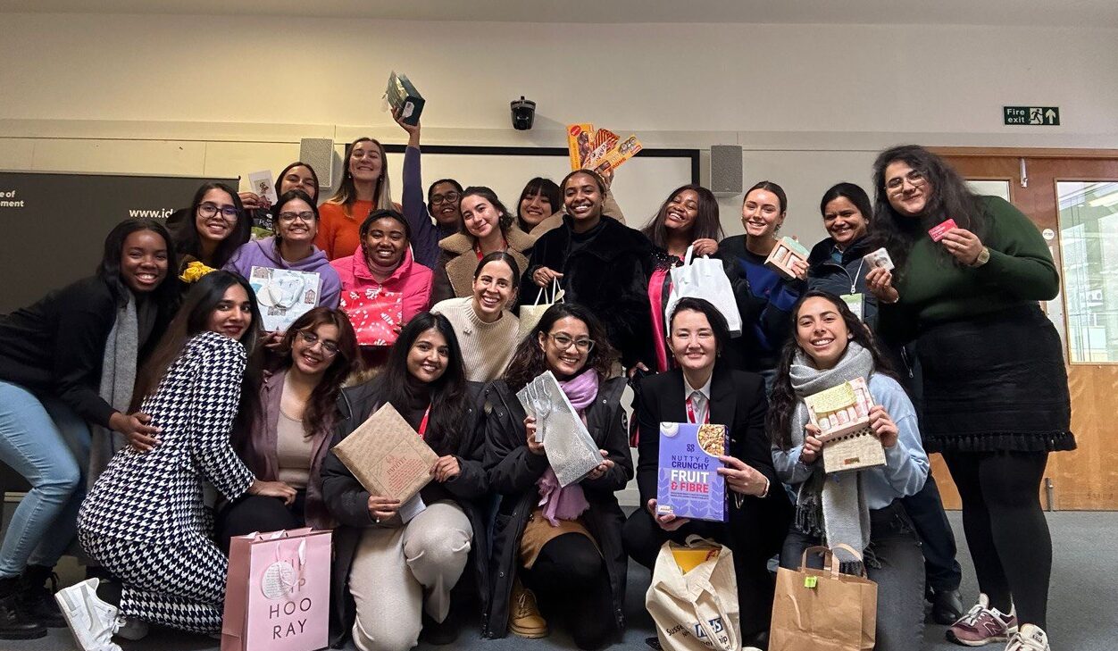 A group of women are gathered together looking at the camera and holding presents that have been exchanged as part of a Secret Santa party.