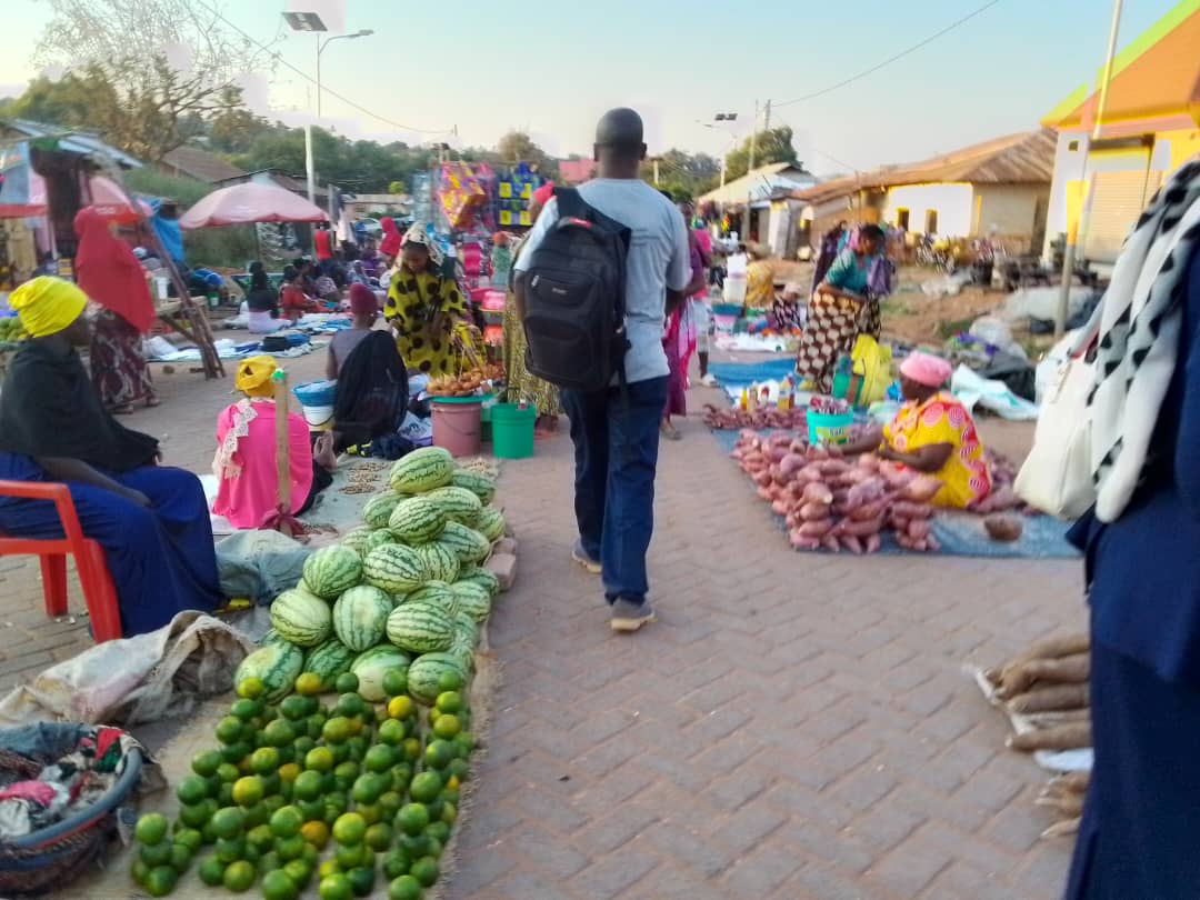 A man walks away from the camera through a busy food market. He is carrying a rucksack over his shoulder. Market vendors have their food laid out on the ground, including watermelon and avocados.