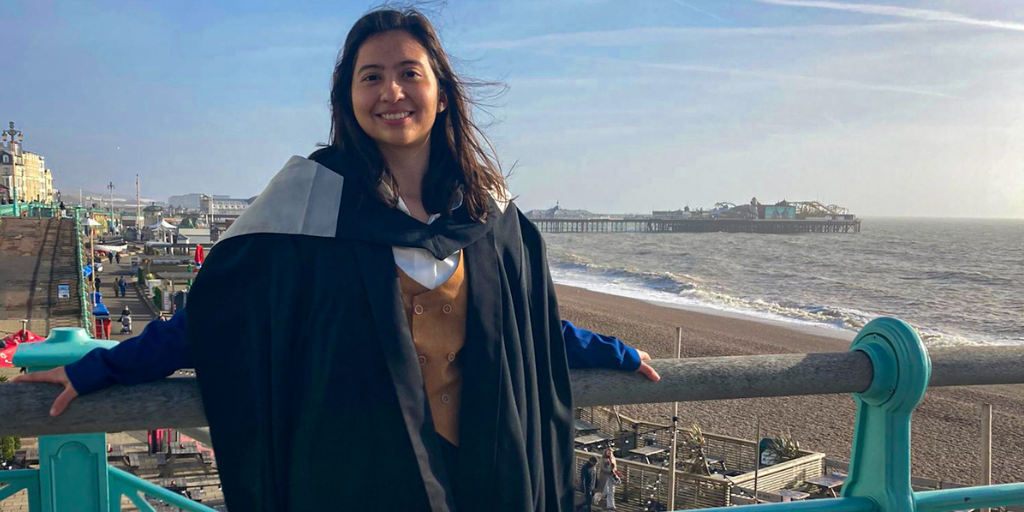 A girl with long dark hair wearing a black graduation gown standing in front of the sea.