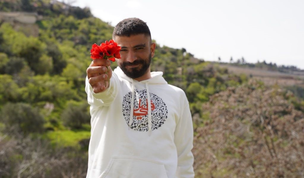 A man wearing a white hooded top smiles at the camera, holding a red flower in their hand. The background shows trees and hillside.