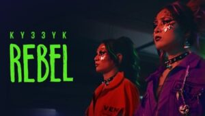 Two girls dressed in punk rock clothes and make up look ahead. The lighting on the girls is red, the background is dark. The title of the track and band name is written in green