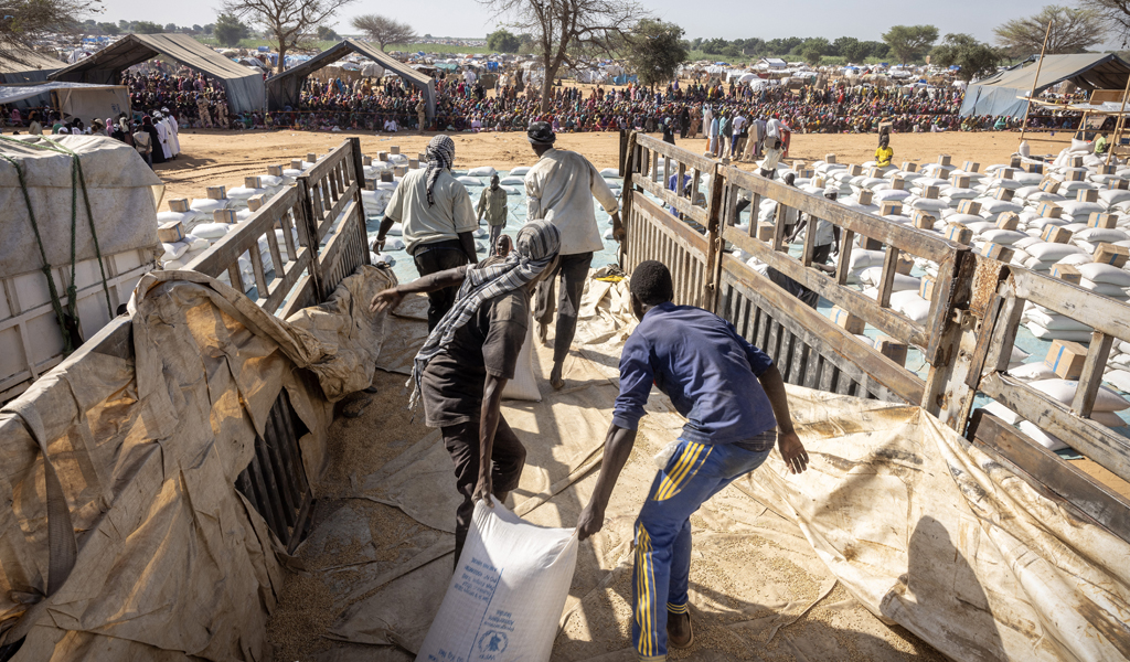 A photo of men dragging bags of food off a lorry. In the distance are a large group of people from Darfur awaiting the food aid.
