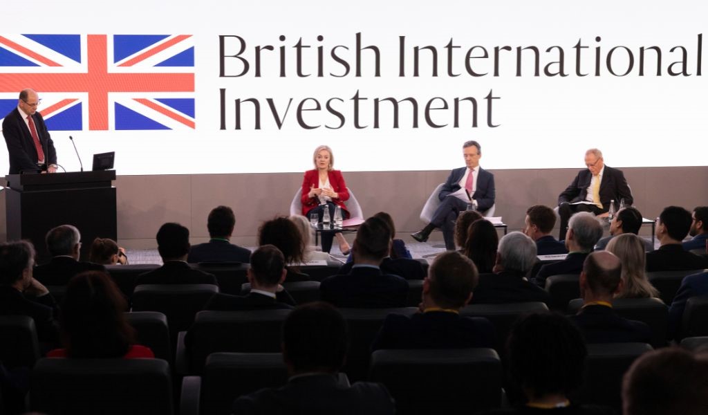 Three people sitting on a smart stage in front of an audience with a large sign behind them that reads 'British International Investment' alongside a union jack flag.