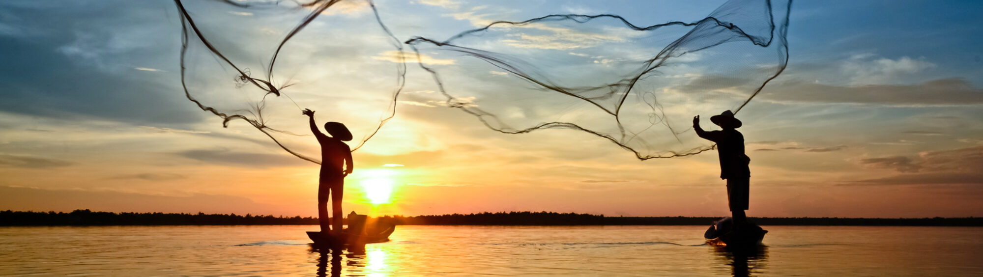 This is a photo of two people casting nets out to sea. Behind them is a sunset.