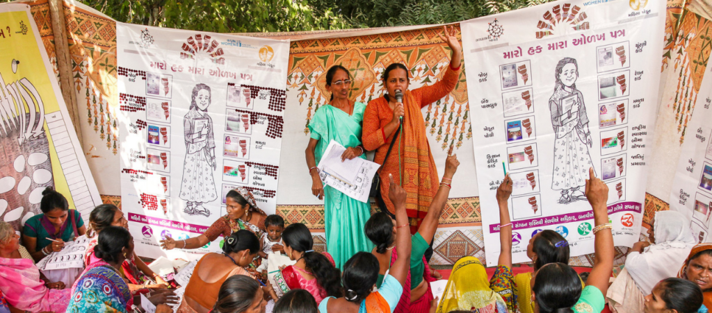 A group of 20 women in saris sitting on the floor, four with their hands in the air. Two women in sarism one holding a microphone, address them. There are posters in the background.
