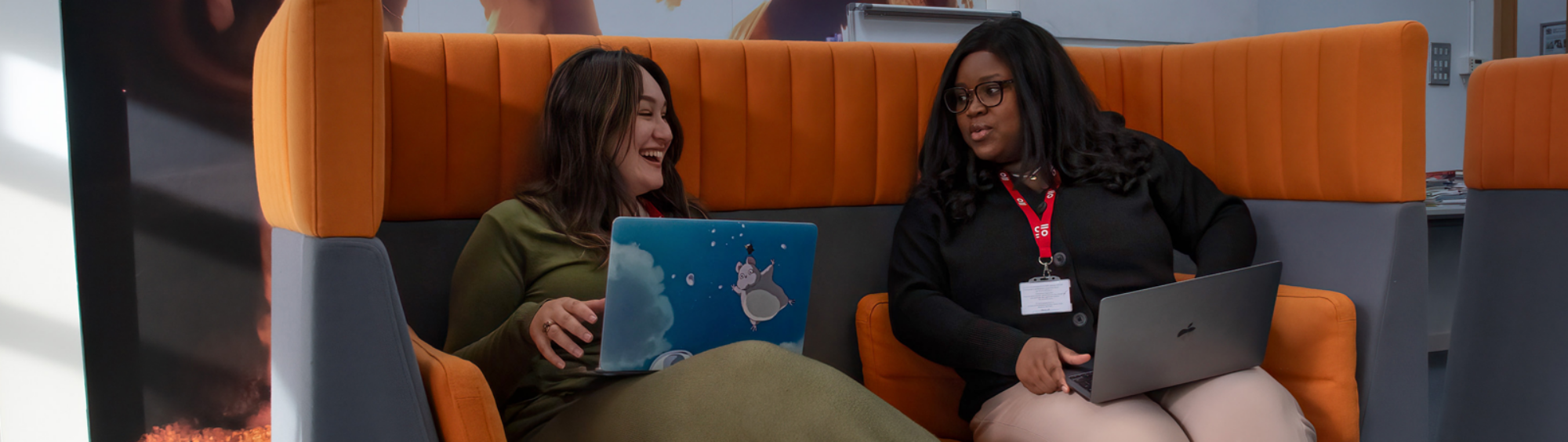 Two students with laptops sit on an orange sofa as they talk to each other.