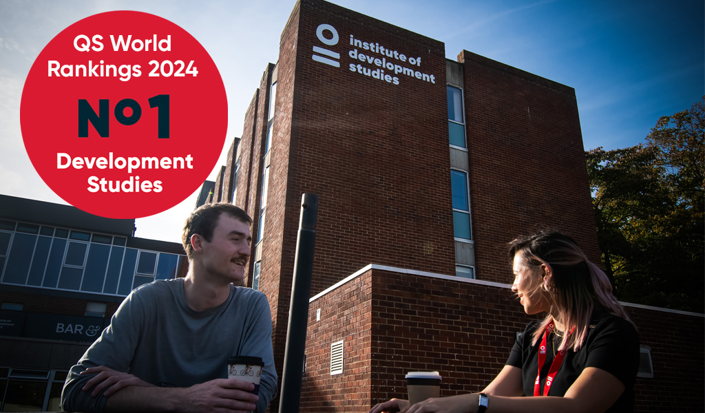 Two people sitting facing each other with cups of hot drinks. They are sitting outside in front of a building with the Institute of Development Studies logo and name written on it.