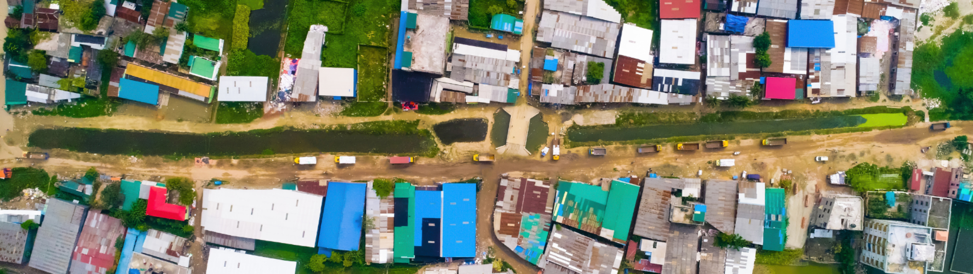 An aerial view of a slum in Bangladesh. The camera is looking down on a series of about 50 dwellings, all different colours, many with corrugated iron roofs.