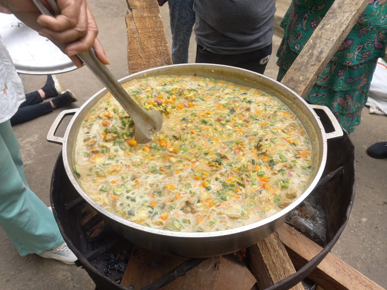 A photo of a stew being prepared. The stew is colourful, orange, yellow and green. It's on an outside cooking stove and someone is stirring it with a big spoon. You can see people standing around.