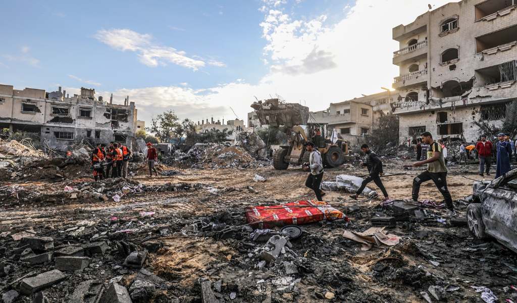 A scene of men walking through rubble of demolished buildings, with heavily damaged buildings still standing in the background. A group of people in orange vests in the background appear to be carrying a stretcher.