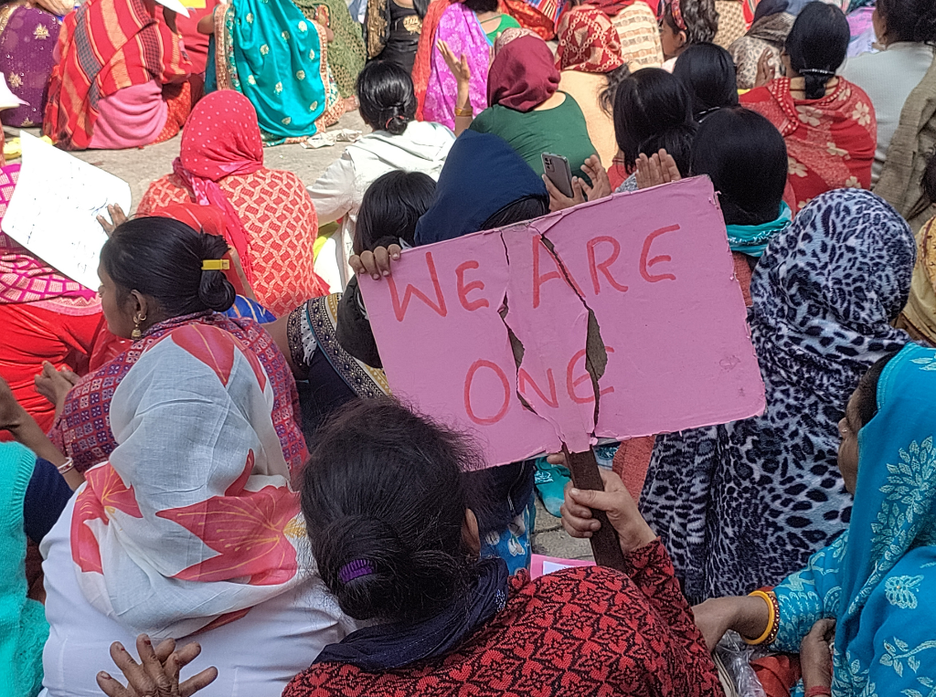 Rows of women with dark hair and their backs to the camera, one of them holds a pink banner that says "we are one". 