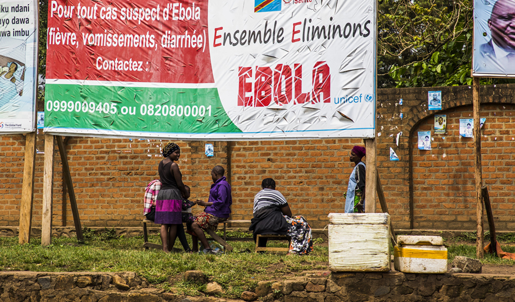 A group of five people, with three sitting and two standing, gathered outside on grass, under a large banner with some information in French and 'EBOLA' in large letters.