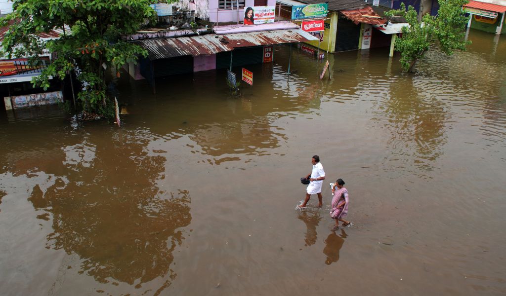 Two people walking through flood water in front of a row of basic shops with corrugated metal roofs.