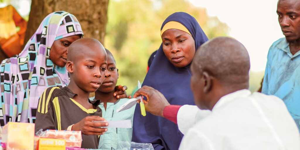 A child is sitting on the lap of a Woman in Nigeria. A male Doctor is holding up a piece of paper to the boy.