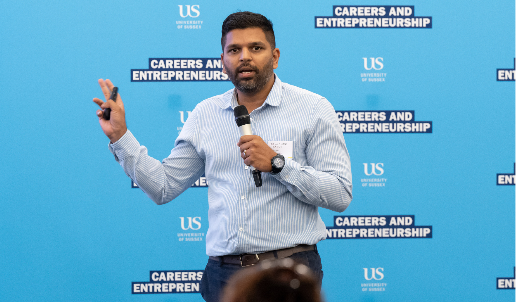 An Indian man with short dark hair, holding a microphone. He stands in front of a blue wall, which has the University of Sussex's logo on it, as well as the lettering 'Careers and entrepeneurship'.