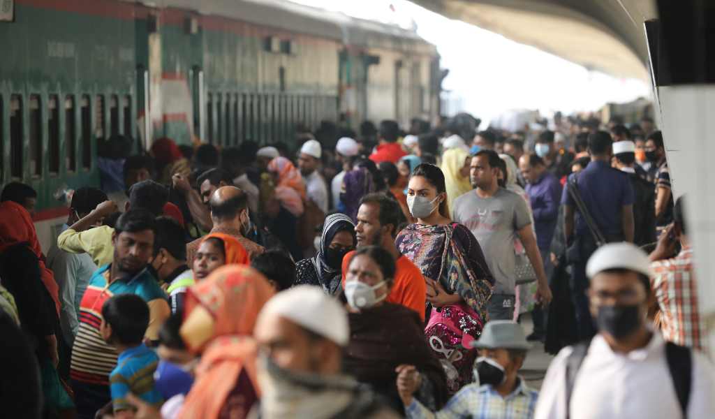 A crowd of people, some wearing face masks, many wearing colourful clothes or headscarves, on a packed train station platform. A train is visible on the left..