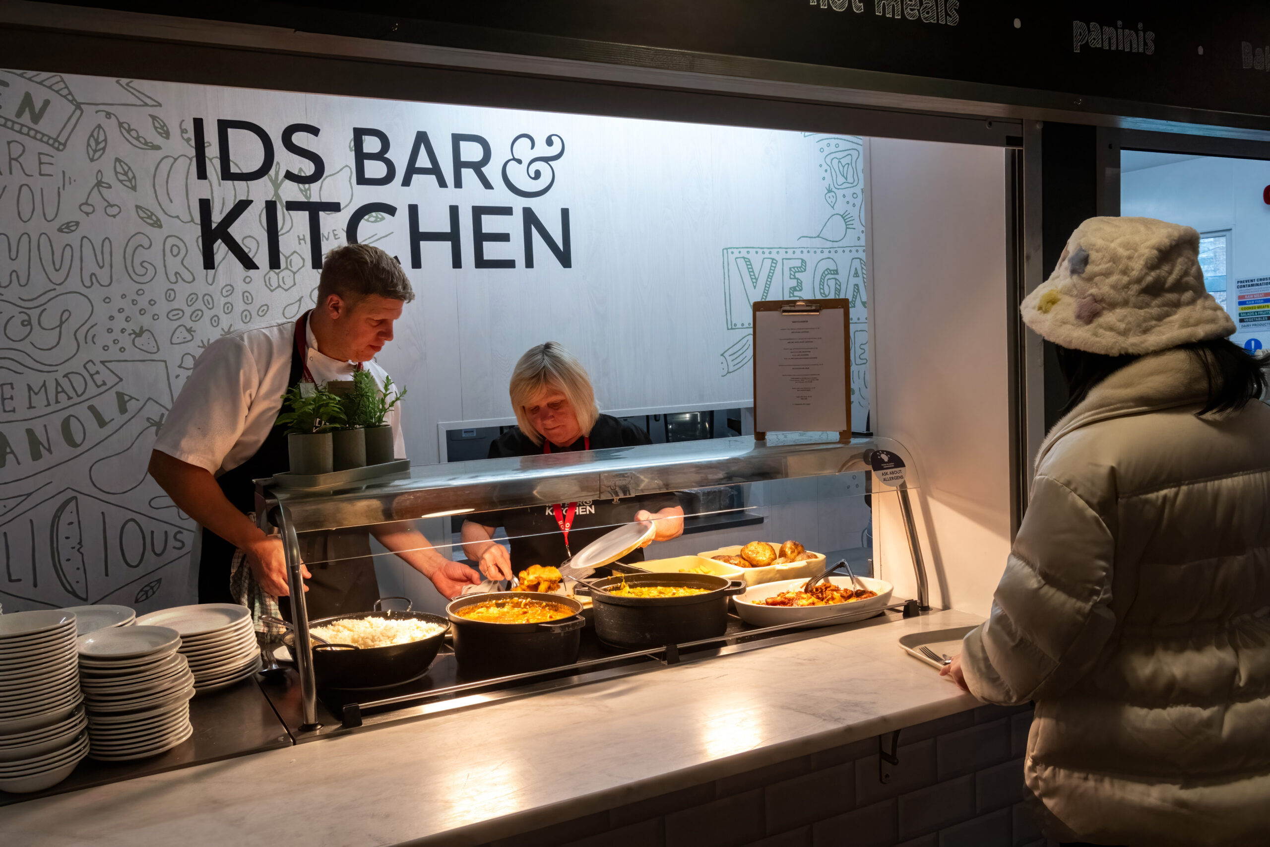 Two people stand behind a counter with food and serve a customer. Behind them is a sign that says 'IDS Bar & Kitchen'