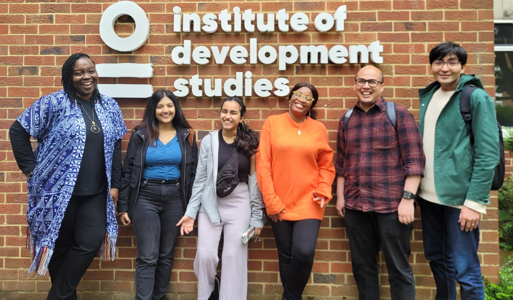 Six people stand against a brick wall, smiling at the camera, in front of the IDS logo in white. They are a mixture of genders and nationalities.