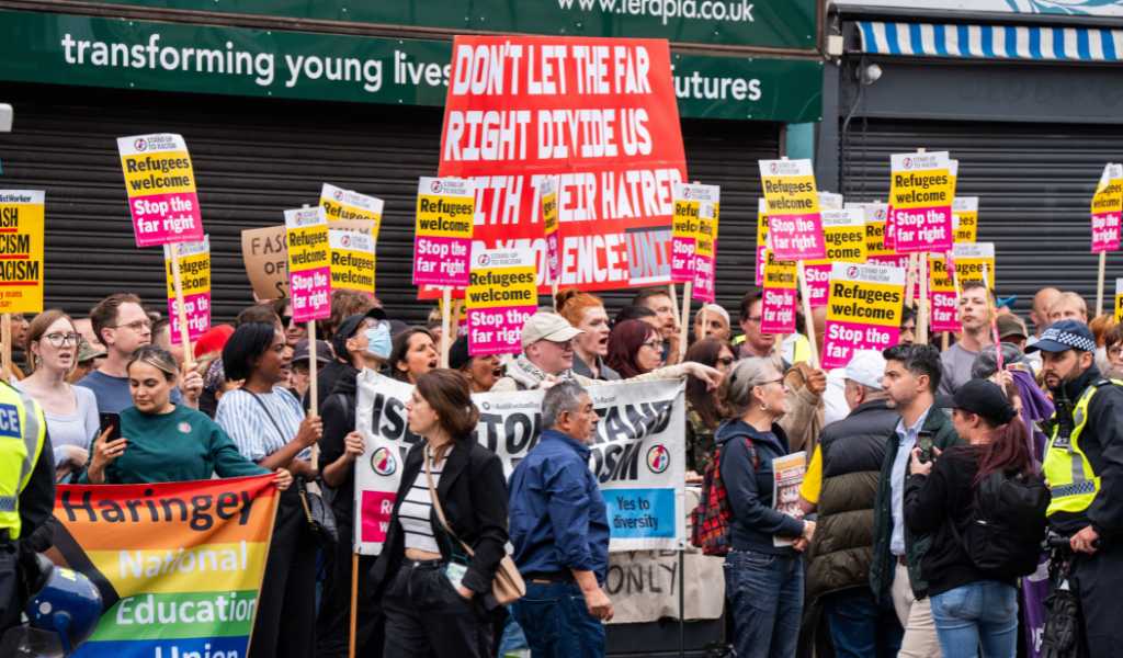 A crowd of people holding up yellow and red banners that read 'Refugees welcome, Stop the far right'. They are standing outside in front of shops with black shutters down.