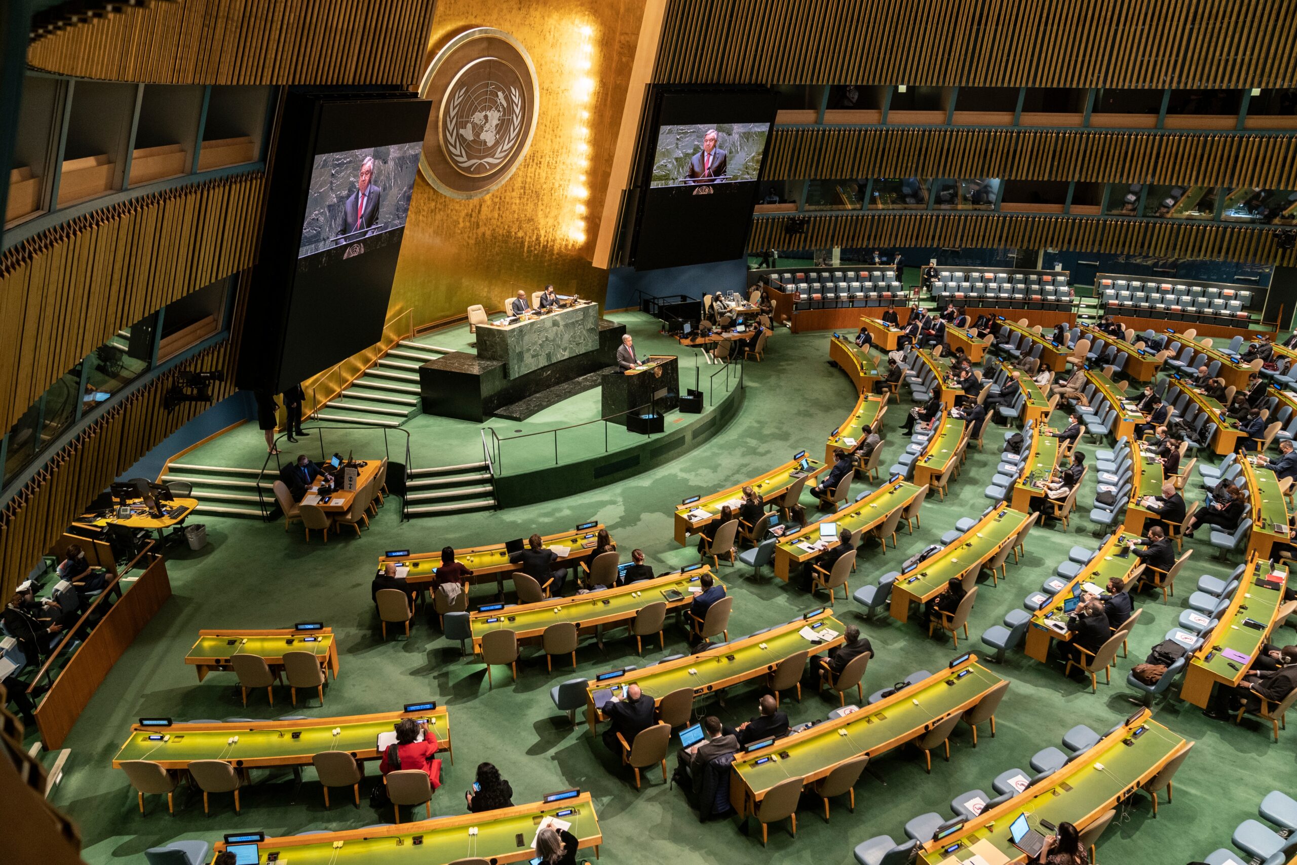A birds-eye view of a UN meeting. There is a table on the stage, with a semi-cirle of tables in front of the stage.