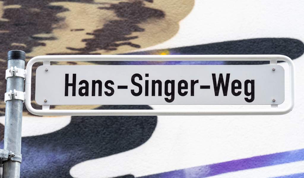 A white metal street with 'Hans-Singer-Weg' written in black letters. White and dark blue abstract shapes from a mural painted are on a wall behind the sign.