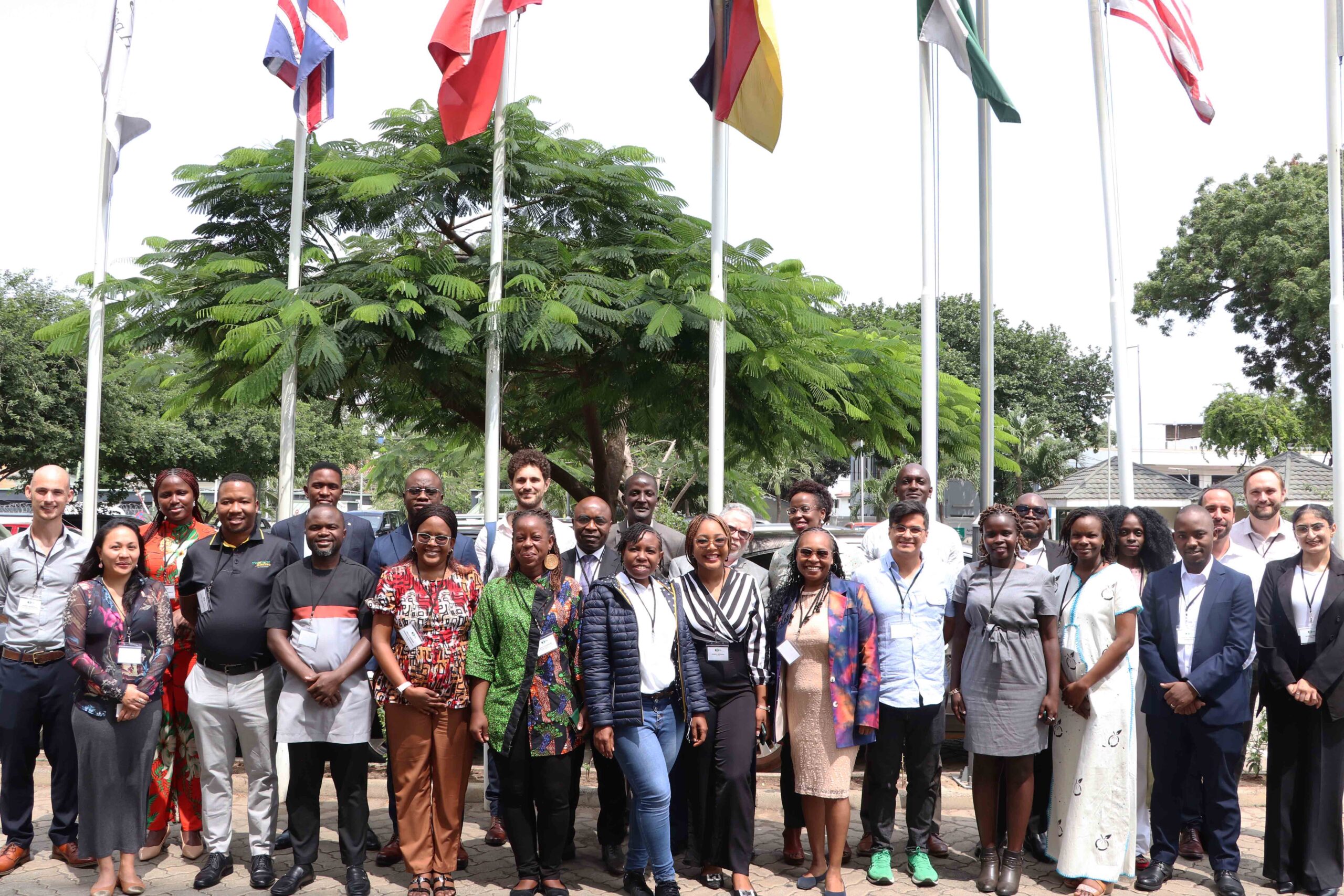 A photo of the ICTD Research on Tax and Development course cohort for 2023-2024 with the in-person module facilitators. There are about 30 adult individuals stand in front of the camera, smiling, outdoors. Behind them are a row of flag poles and trees. 