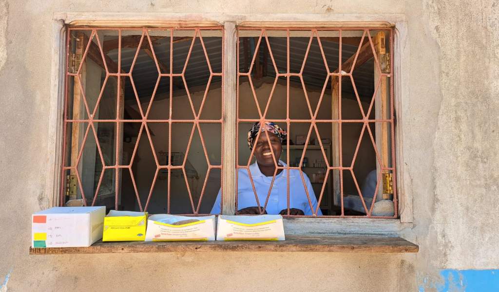 A smiling woman wearing a headscarf and blue shirt standing behind a window covered with metal bars in a diamond shape. Four shallow white and yellow cardboard boxes sit on the windowsill.