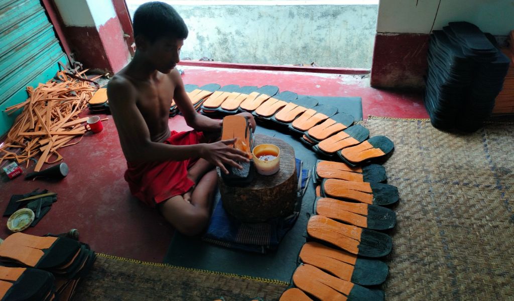 A boy sitting cross legged on a hard floor putting glue on an orange and black coloured leather sole with his fingers. Piles of soles for shoes are on the floor in front of him in a semi circle.