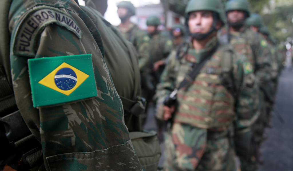 A photo of a Brazil flag patch on the arm of a Military Uniform. In the distance are a group of male Brazilian soldiers