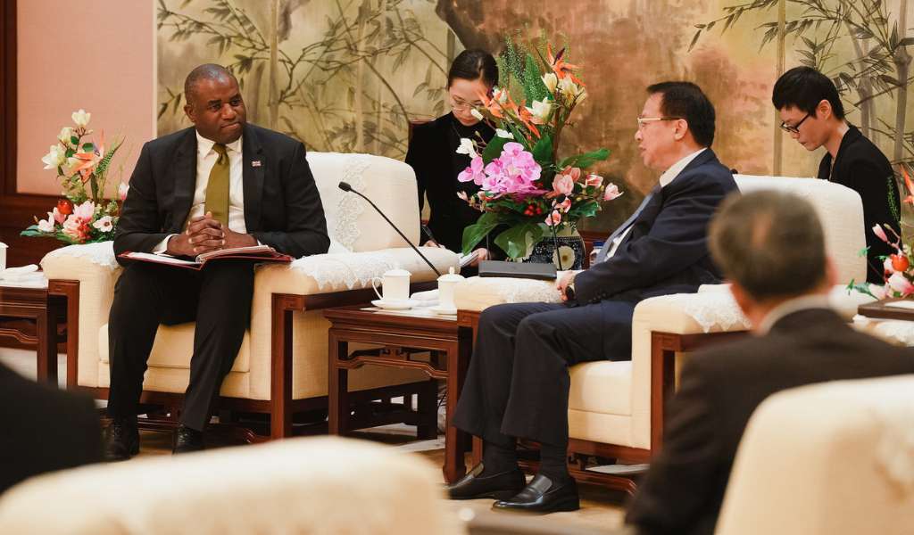 Two men in suits sitting on large white chairs, facing each other, in a formal setting. Two women in black sit discreetly behind them.
