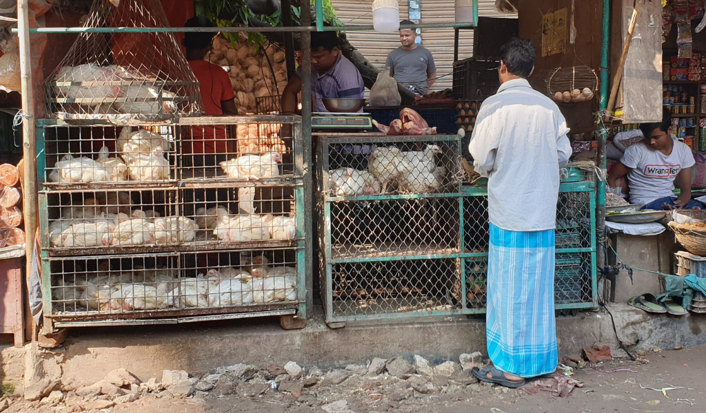 A man selling chicken behind the counter to a man in a white shirt and blue dhoti in Dhaka, Bangladesh.