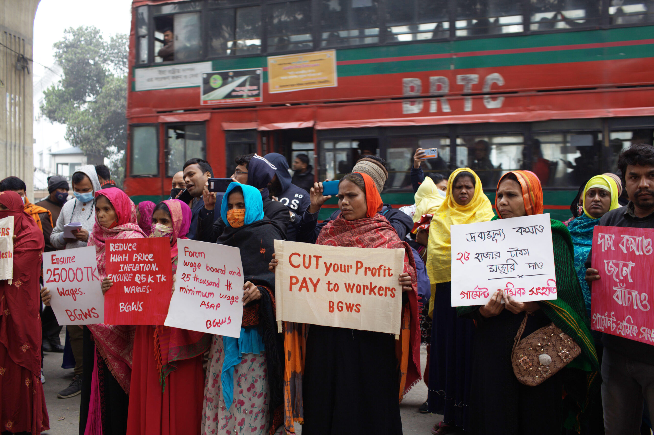 A group of women stand outside holding placards protesting to raise the minimum wage for garment workers. Behind them, a red bus goes by.